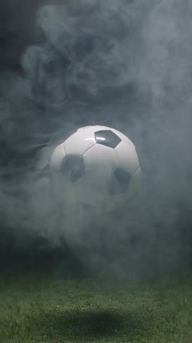 A soccer ball hovers in midair above a grassy field at night with white smoke drifting across the atmosphere