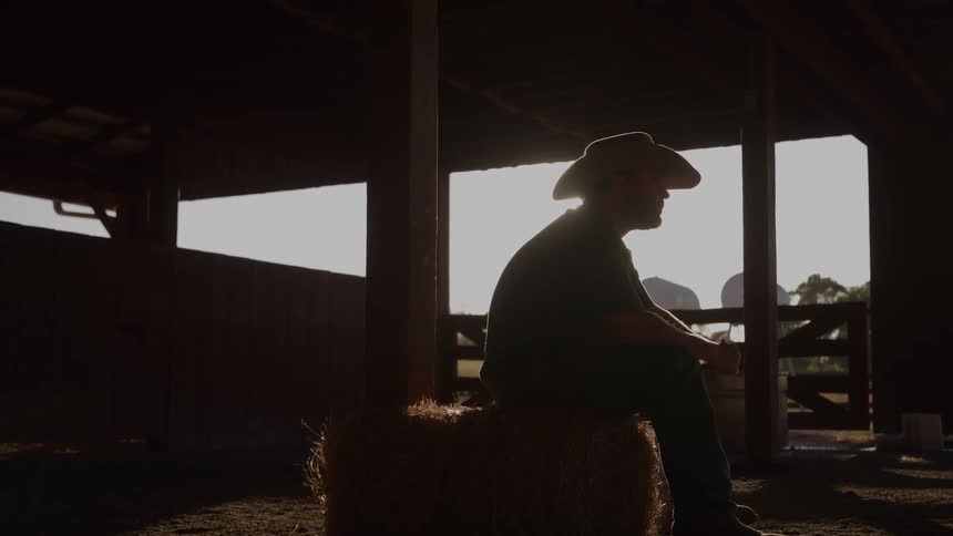 A strong silhouette of a person in a cowboy hat is captured sitting on a hay bale inside a dark barn or shed the setting sun blazes through the doorway creating a dramatic halo effect and framing the figure in golden light