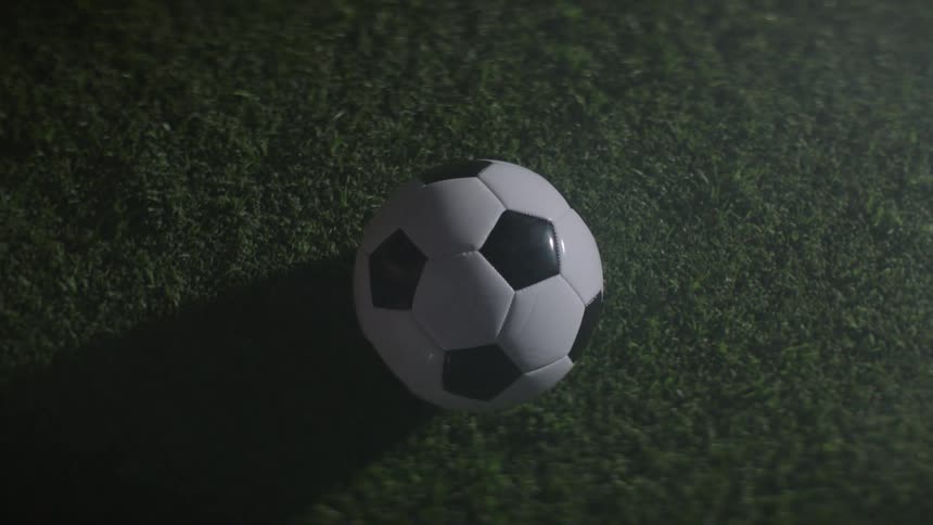 A soccer ball appears from the dark top of the frame descends and lands softly in the center of the shot onto a patch of short green artificial grass before resting completely still