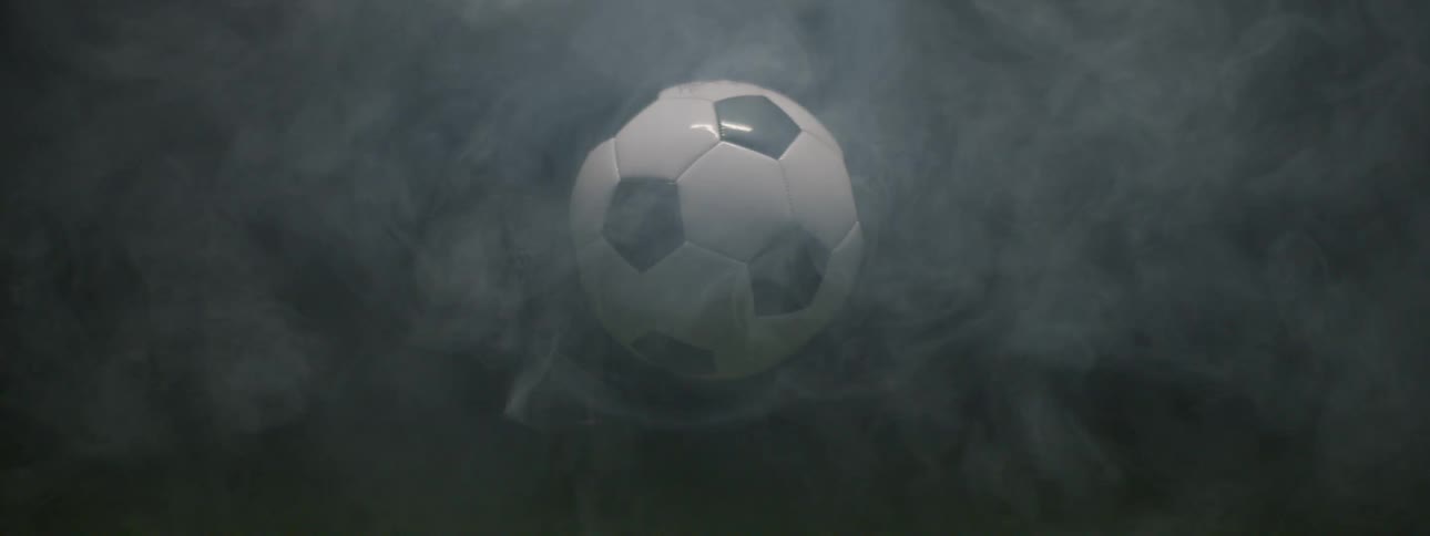 A closeup shot of a patch of green grass turf transitions into a dark smoky environment as a black and white panel soccer ball descends and stabilizes hanging suspended midair