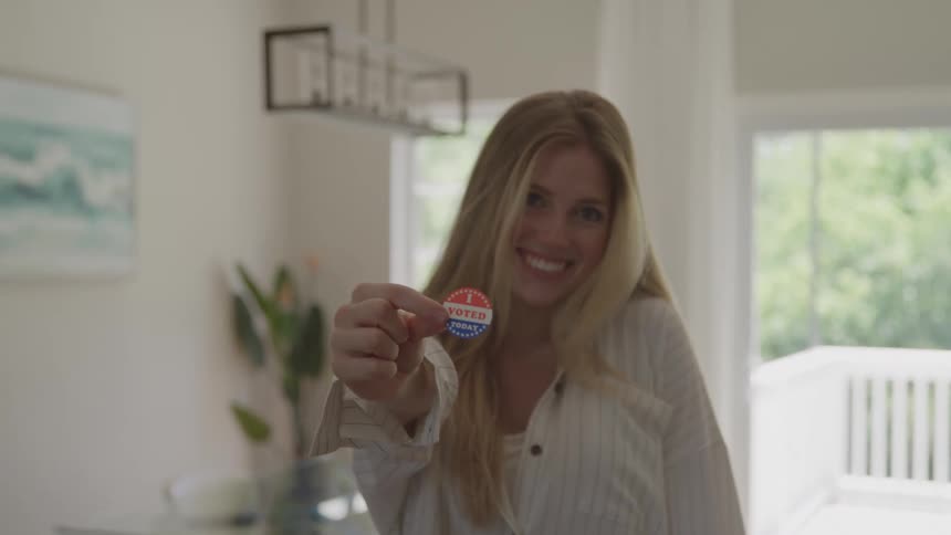 A young woman with long blonde hair stands in a brightly lit room and smiles directly at the camera while holding up and presenting an i voted today sticker in her right hand towards the lens