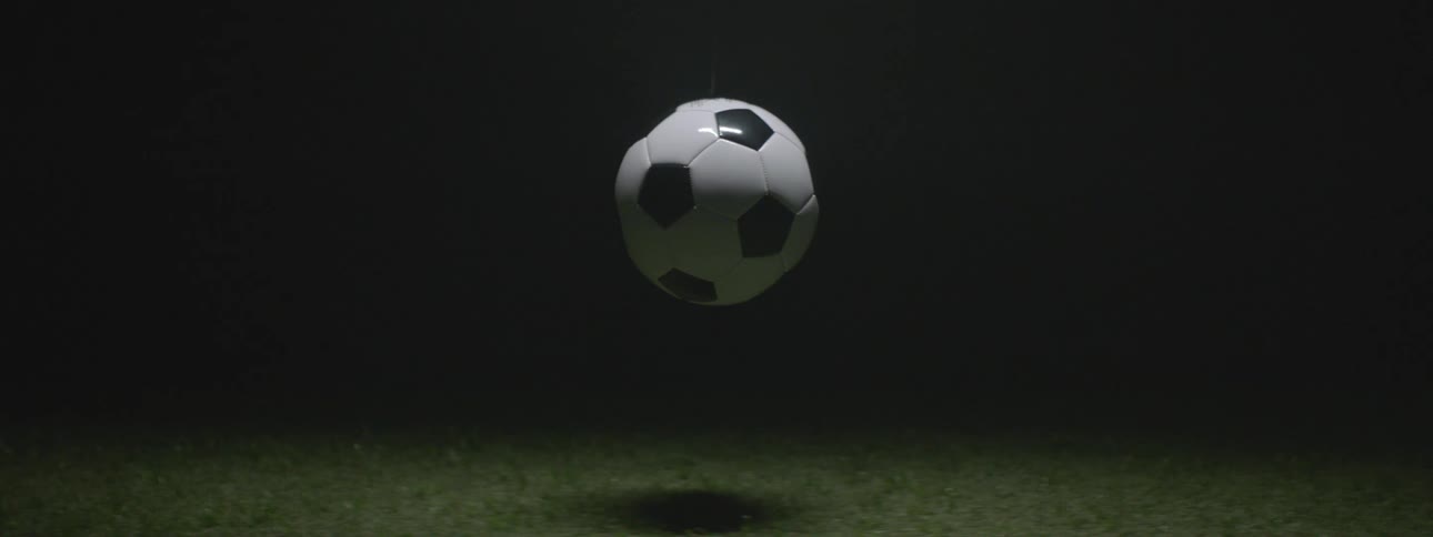 This clip features a traditional black and white soccer ball hovering slightly above a patch of grassthe setting is dimly lit with the soccer ball being the central slightly illuminated object against a dark background casting a soft shadow on the g