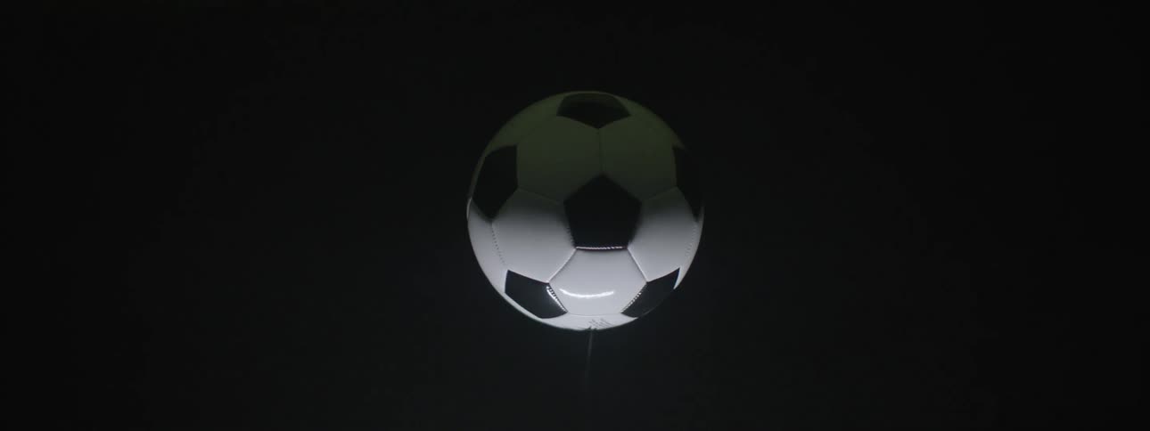 This is a closeup shot of a classic black and white soccer ball football slowly spinning while suspended on a thin string or wire against a dark background the right side of the frame shows a faint hint of green suggesting it is near a grass surface