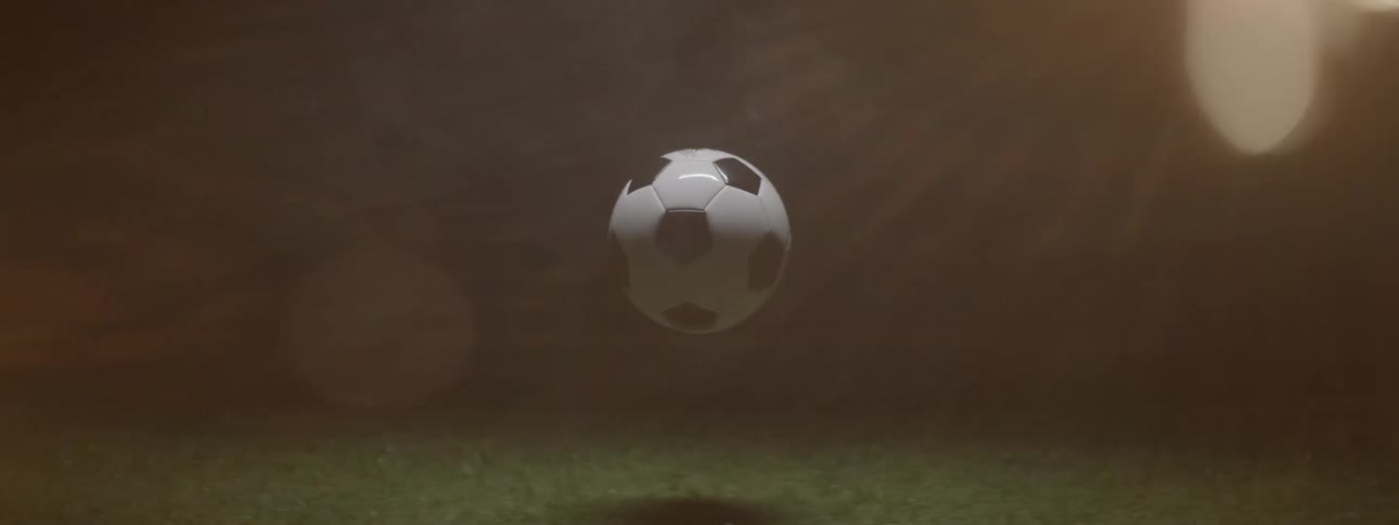 A black and white soccer ball floats in the air illuminated from above and reflecting on a grassy ground in a dark atmospheric setting