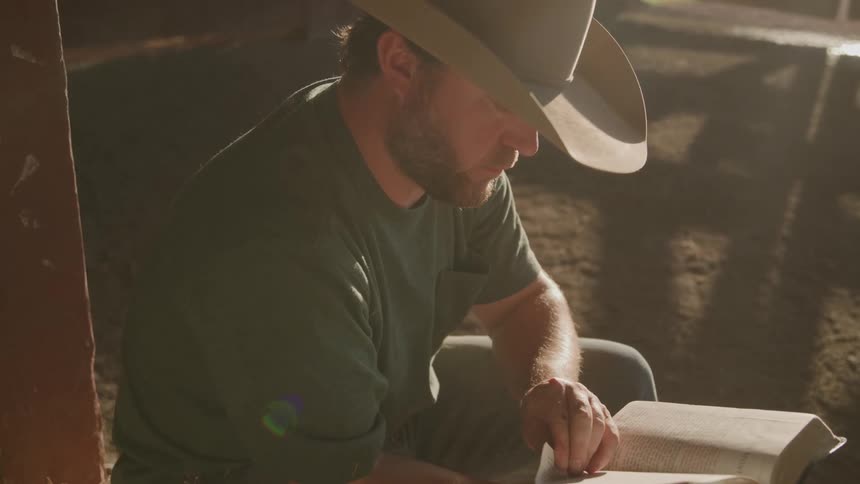 Person wearing cowboy hat reading a book indoors softly lit by sunlight