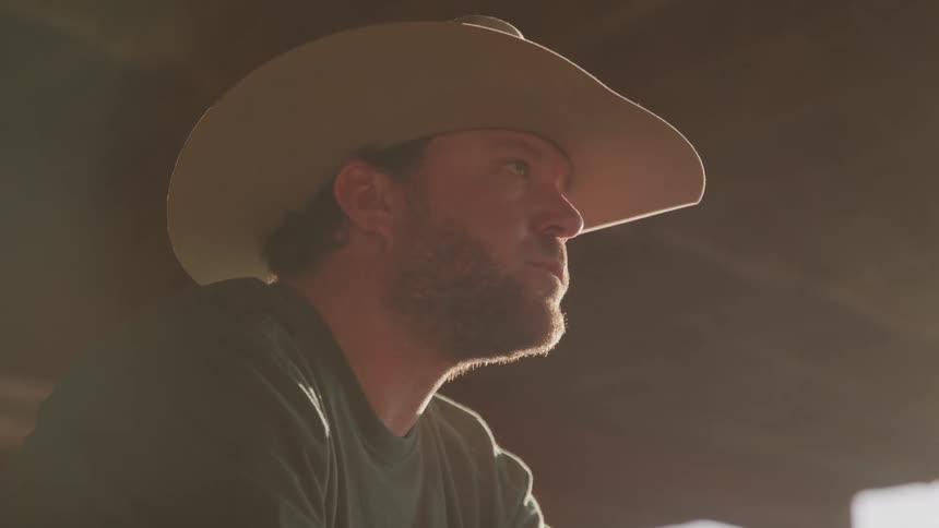 A close side view of a bearded man in a cowboy hat looking up in a dim space with bright sunlight shining under the brim of his hat