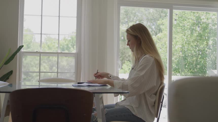 Young woman wearing a white shirt and jeans sitting down on a chair in front of a wooden table reading and writing on sheets of paper indoors during day time