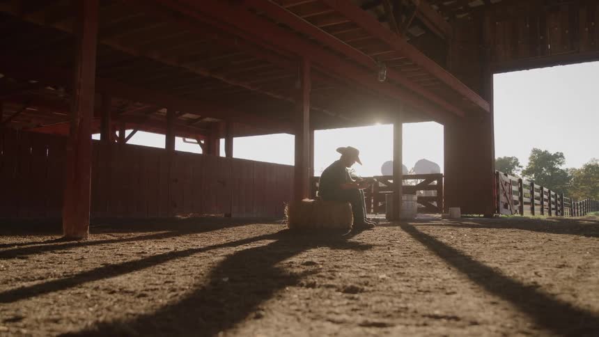 Person in cowboy hat sits on hay bale contemplating something in hands inside sunlit barn
