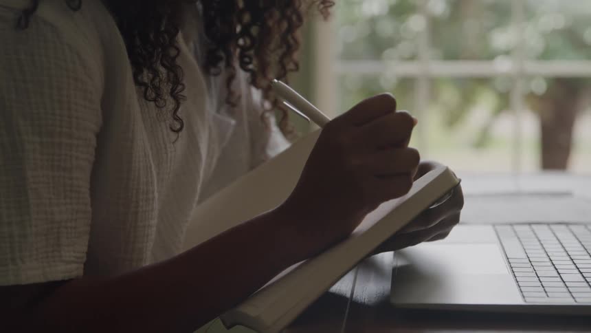 The video shows a closeup of a person likely a woman with curly hair sitting indoors near a window she is wearing a white shirt and is seen writing in a small notebook with a white pen a laptop is visible on the desk to her right