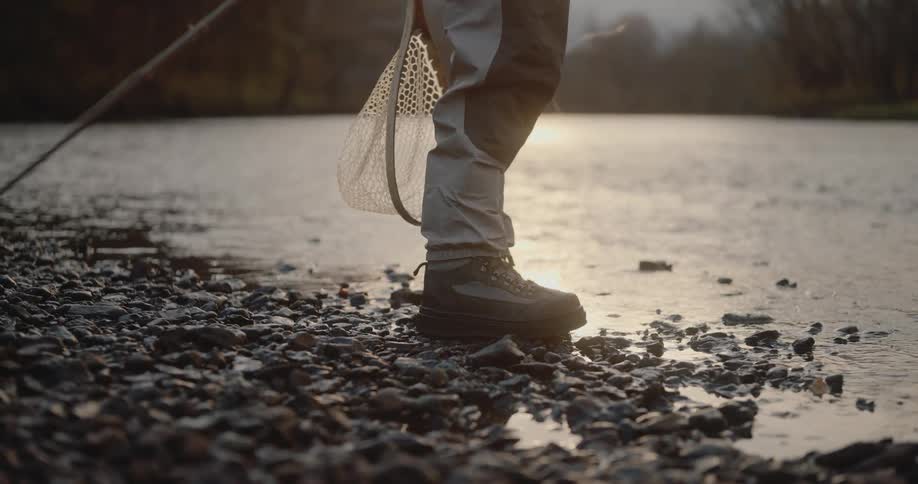 This video shows a closeup of a persons legs wearing waders and sturdy fishing boots standing on a rocky bank next to a body of water they are holding a fishing rod and a landing net in their hand suggesting they are fly fishing or angling