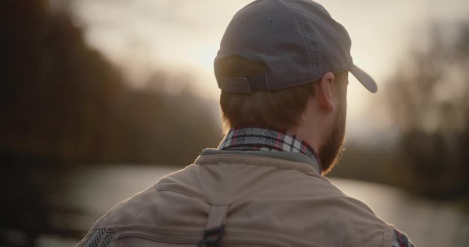 A man with a beard and baseball cap stands outdoors fishing with his back to the camera looking towards a bright sunset over water and trees