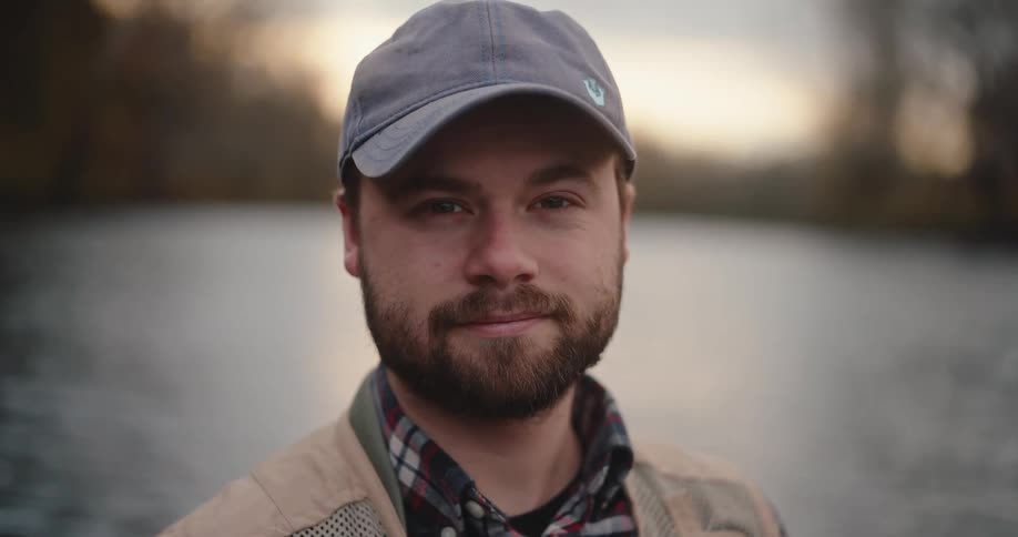 A close view of a smiling man with a beard wearing a baseball cap and a plaid shirt outdoors by a body of water on an overcast day