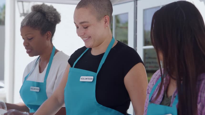 Three women with volunteer badges smile while helping at an outdoor community event