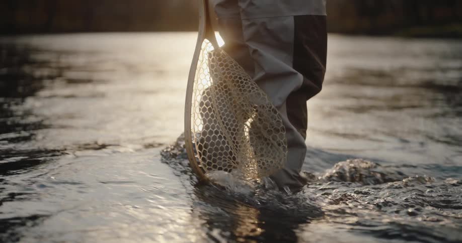 A low angle close view of a person wearing gray fishing waders standing in a sunlit river holding a net in the flowing water
