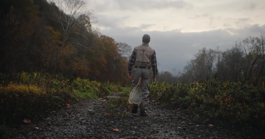 A man dressed in outdoor gear including a vest and waders walking away down a dark wet gravel path and carrying a fishing net in his hand moving toward a valley landscape with muted autumn foliage under a overcast sky