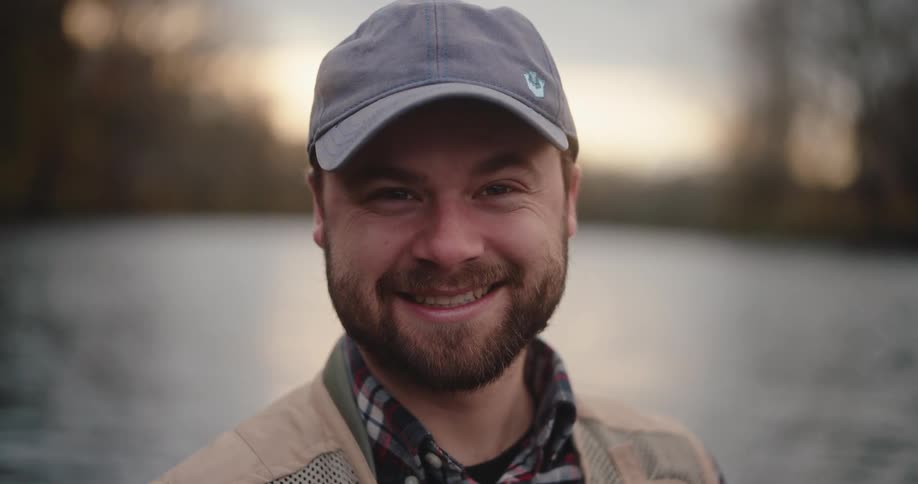 Bearded man wearing a baseball cap standing in front of a river and smiling outdoors during day time