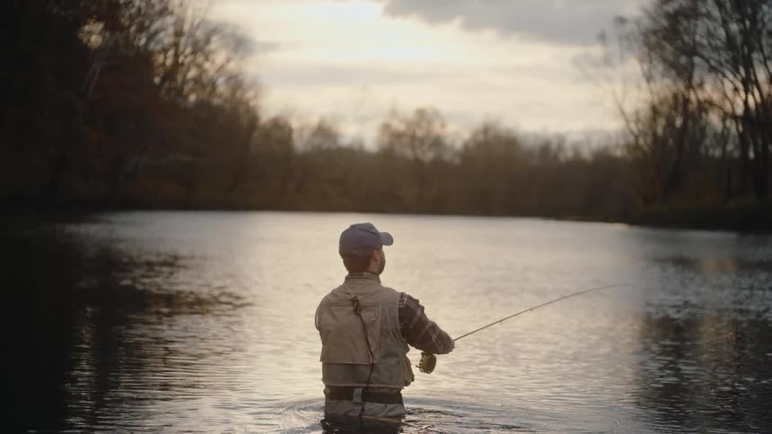 Young man wearing a baseball cap and a vest fishing on a pond surrounded by trees outdoors during dusk or dawn