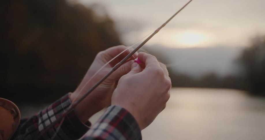 Hands adjust a fishing line on a rod by the river at sunset