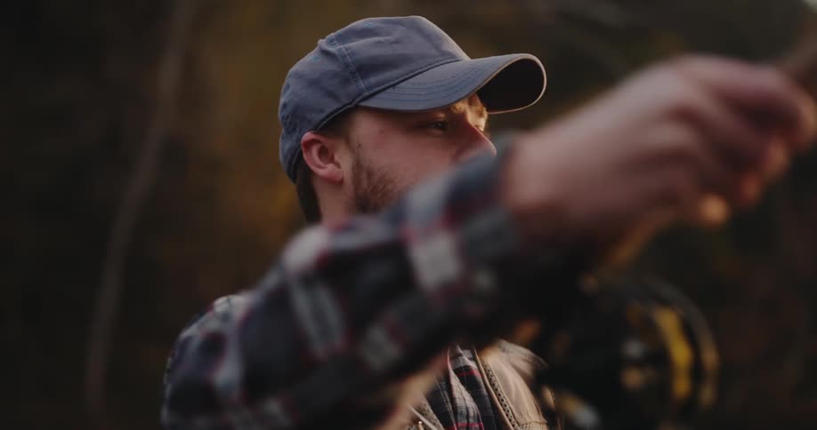A bearded man wearing a baseball cap a plaid shirt and a fishing vest raising his arm to cast a fishing line with his face illuminated by a warm sidelight and the background consisting of dark blurred foliage