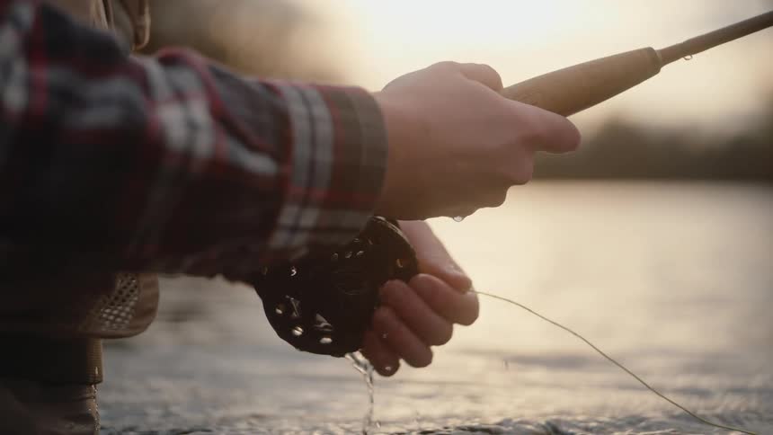 The hands of an angler holding a fly fishing rod and reel while standing in shallow water with the reel dripping water as the persons other hand momentarily adjusts the line and releases it over the water surface during sunset