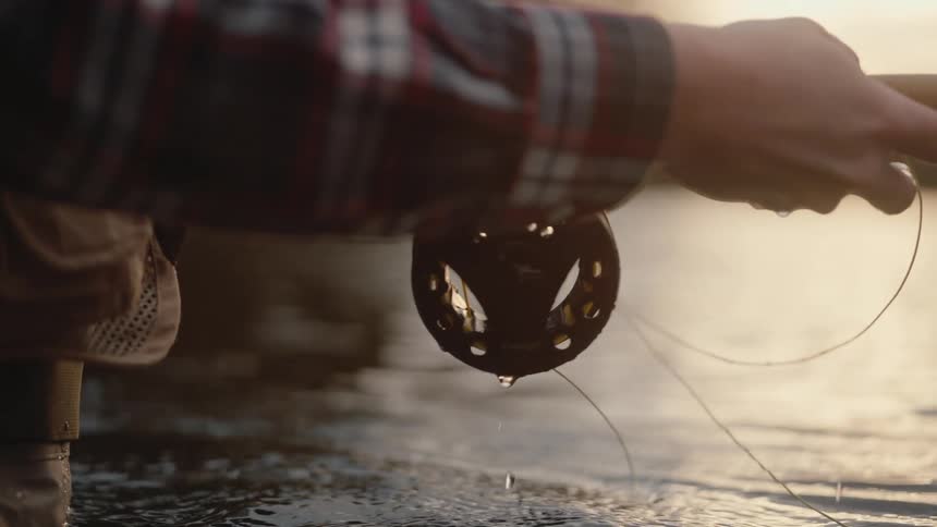 Person in plaid shirt and waders holding fly fishing rod with water droplets on reel