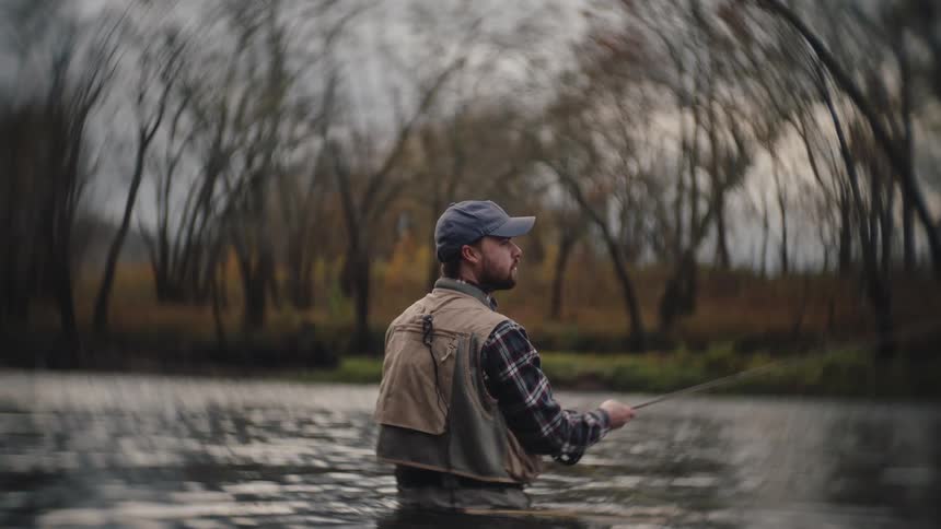 A man with a baseball cap and fishing vest is looking forward while standing in a river against a tree lined background