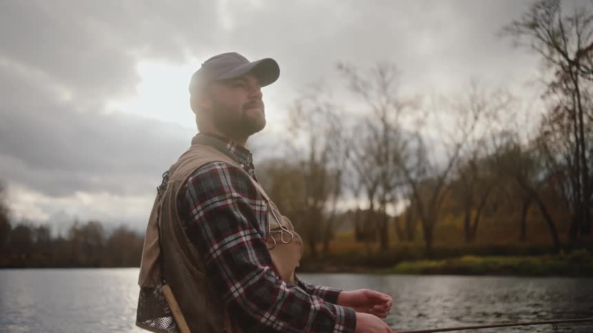 A man with a beard wearing a dark baseball cap a plaid shirt and a fishing vest standing by a body of water and holding a fishing rod with the scene backlight by the sun diffused behind overcast clouds and surrounded by trees with bare branches