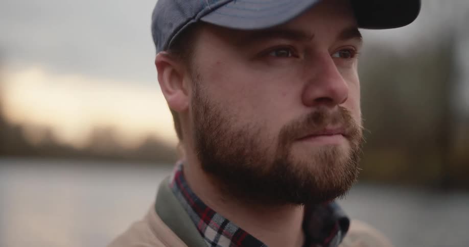 Bearded man in blue cap and plaid shirt under beige jacket facing treelined landscape