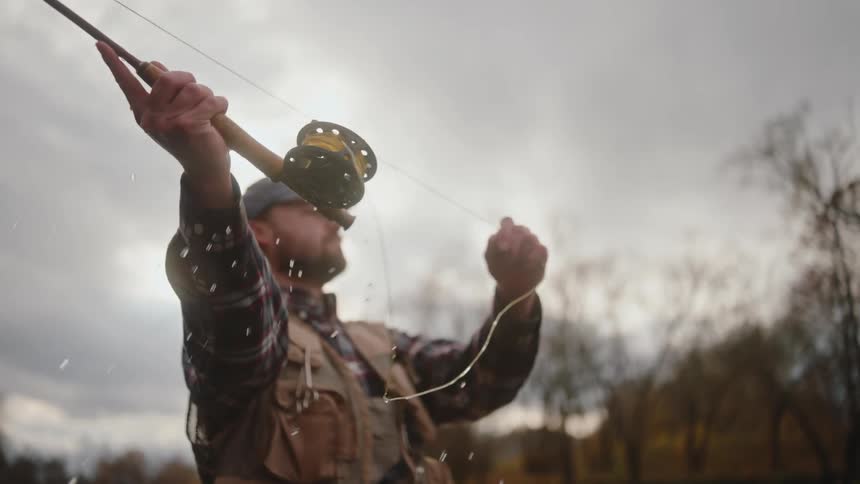 A man wearing a plaid shirt fishing vest and baseball cap is seen from a low angle executing a vigorous cast with a fly fishing rodwater droplets fly off the line and reel as he brings the rod back and then forward against the bright overcast sky