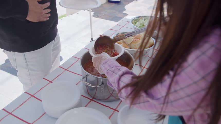 A person serves stew into a paper bowl over a red and white checkered table