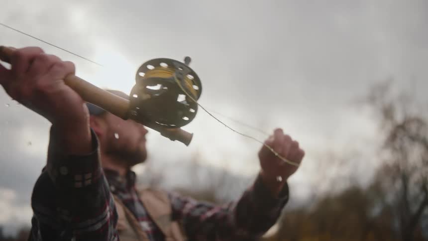 A man with facial hair stands in the river at midday swinging his fishing line across the water