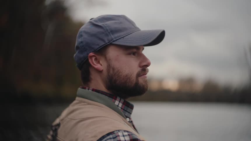 A young man with a beard wearing a grey baseball cap a plaid shirt and a tan vest standing motionless and looking intently offcamera toward a dark blurred body of water and the dense dark foliage on the far bank under an overcast sky