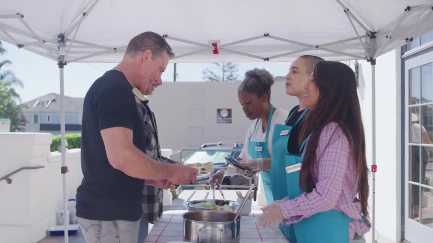 A medium shot under a white canopy tent captures a food distribution line where three female volunteers wearing blue aprons and gloves are serving meals from metal containers onto plates held by two men with a bright sunny outdoor setting