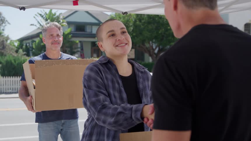 A young woman with a shaved head and a plaid shirt stands under a white canopy smiling as she shake hands with a man in a black tshirt while a second man stands behind them holding a large cardboard box on a suburban street