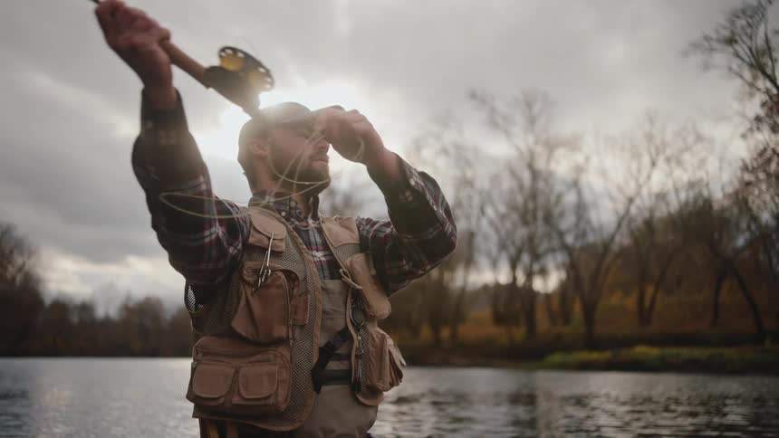 A man fly fishes in a river under a cloudy sky with trees in the background
