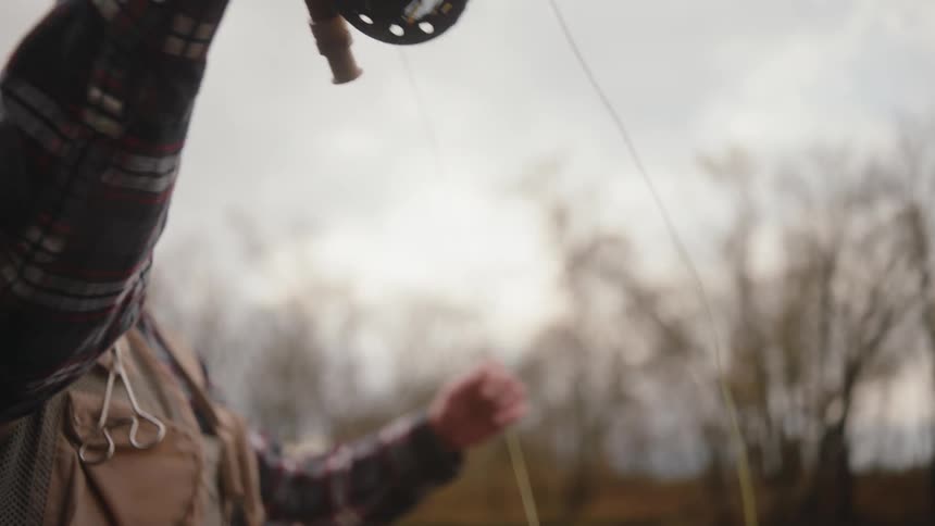 A man wearing a baseball cap and plaid shirt expertly casts a fly fishing rod with a reel outdoors on a cloudy day