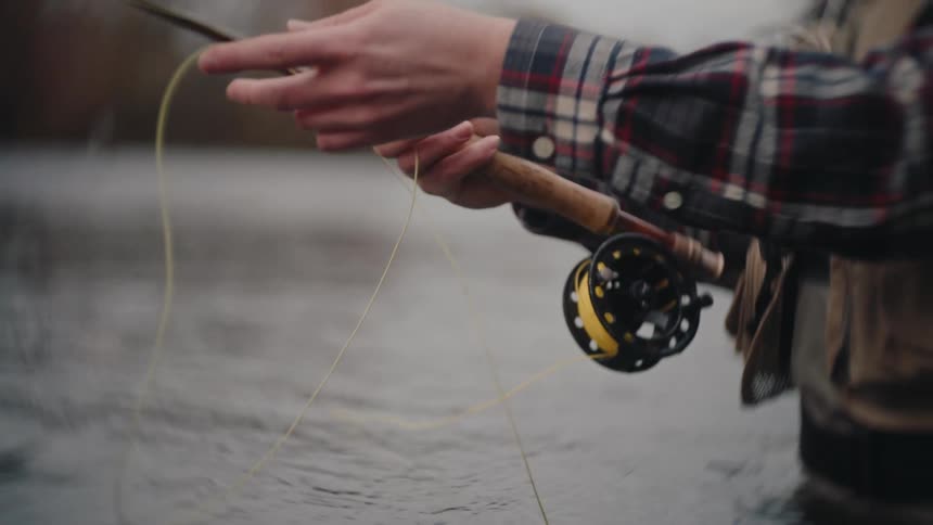 This video provides a closeup of a person fly fishing standing waistdeep in a flowing river or stream the individual is actively casting and managing the yellow fly line with their hands using a rod with a black and yellow reel