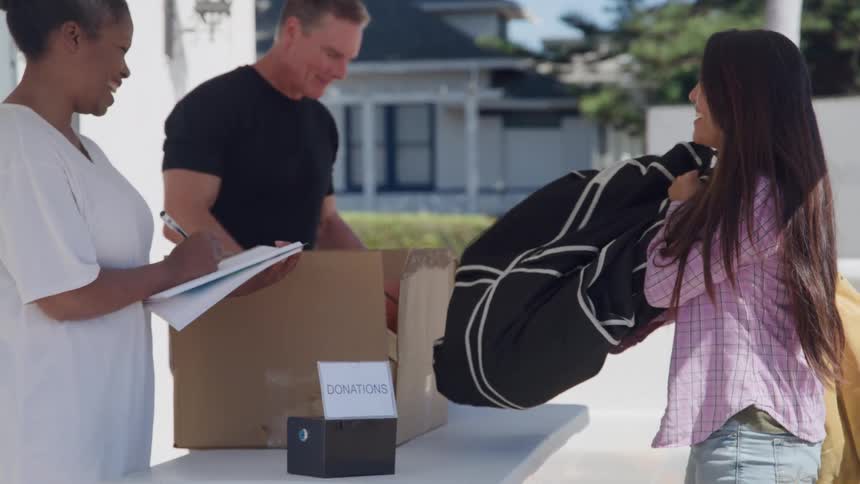 A young woman makes a clothing donation to a diverse volunteer couple managing a donation box and clipboard outdoors on a sunny day