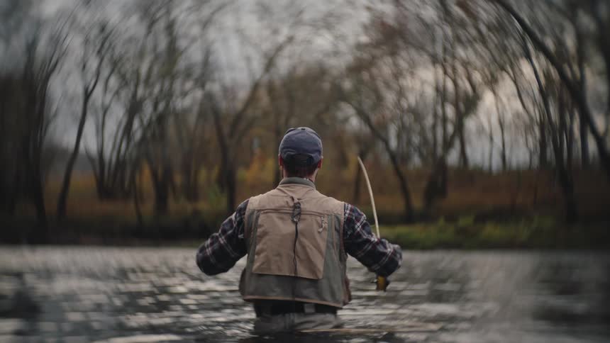 This video shows a person fly fishing standing waistdeep in the murky water of a river or streamthe person viewed from behind is wearing a fishing vest and waders casting the rod with a dense leafless forest forming an arched backdrop