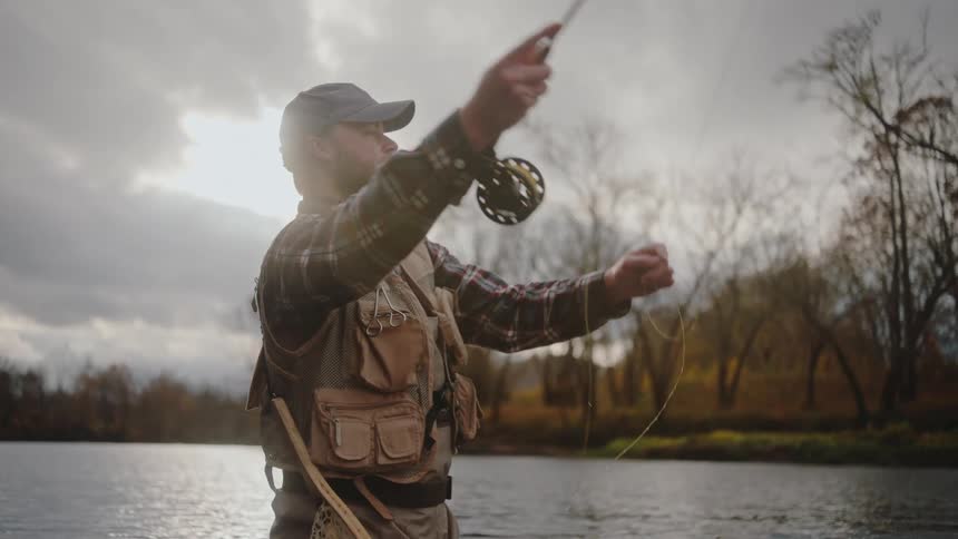 A man in a fishing vest casts a fly rod into a river with autumn trees in the background