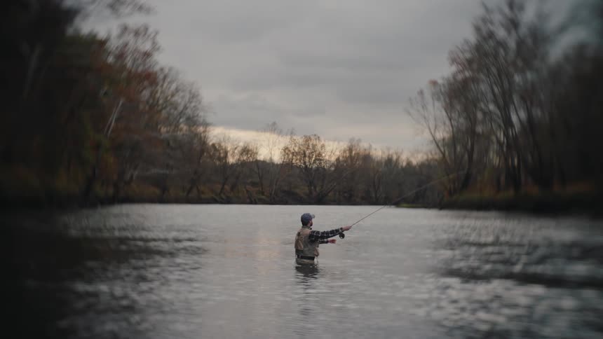 A middleaged man stands waistdeep in the river at midday casting his fishing line into the water