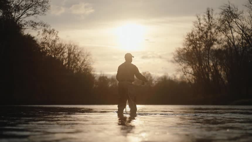 A man in waders fly fishes in a river casting his line into the bright sun reflecting off the water surrounded by a silhouette of bare trees
