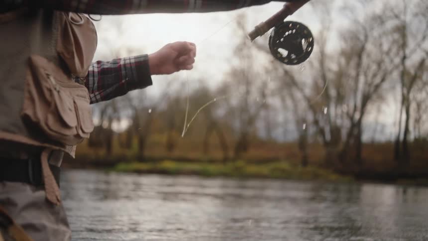 Person in plaid shirt fly fishes in calm river surrounded by leafless trees