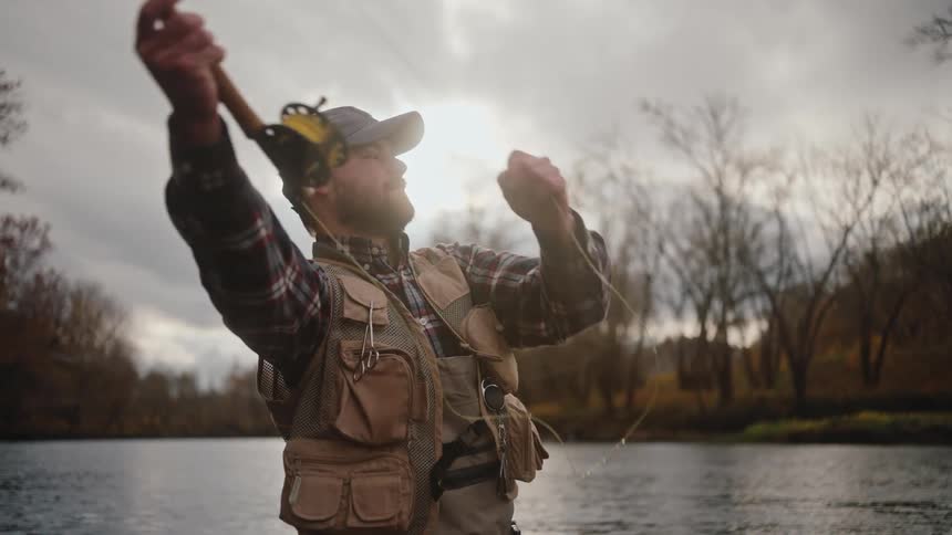 This video shows a bearded man wearing a baseball cap a plaid shirt and a fishing vest standing by a body of water likely a riverhe is fly fishing casting his rod and then holding it while looking toward the water and the wooded landscape under a c