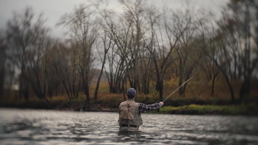Fisherman stands in the river casting a fishing line in front of bare trees