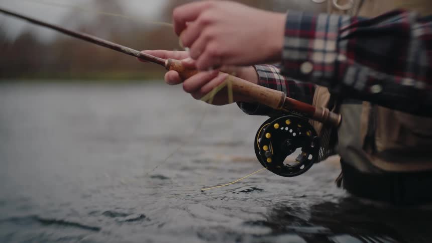 This closeup video shows a person fly fishing standing waistdeep in water and holding a fishing rod with a reel they are wearing a plaid shirt and what appears to be a fishing vest or wadersthe person is actively stripping or handling the line wit