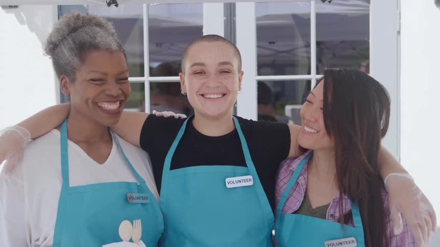 Three women of different races and ages smiling and standing together with their arms around each other wearing a blue apron with a volunteer tag on it outdoors during day time