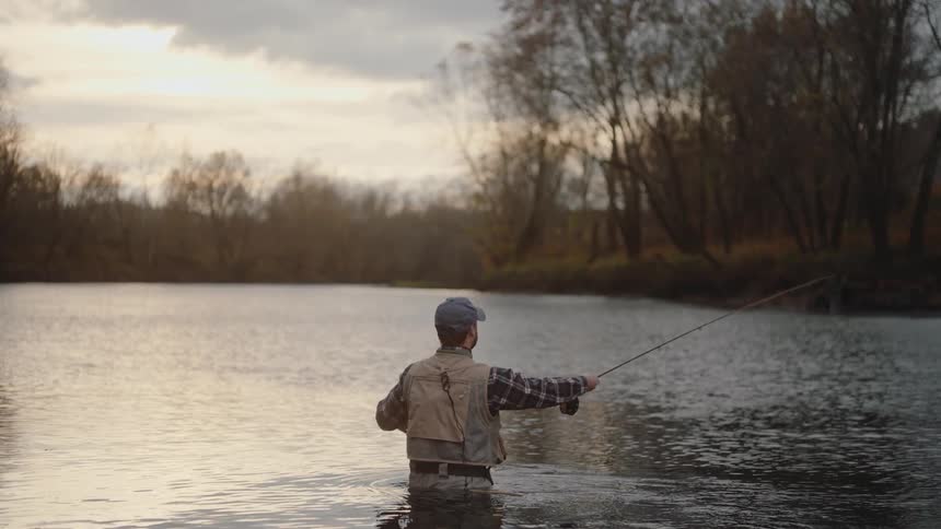 Person fishing in shallow river with leafless trees in the background under cloudy sky