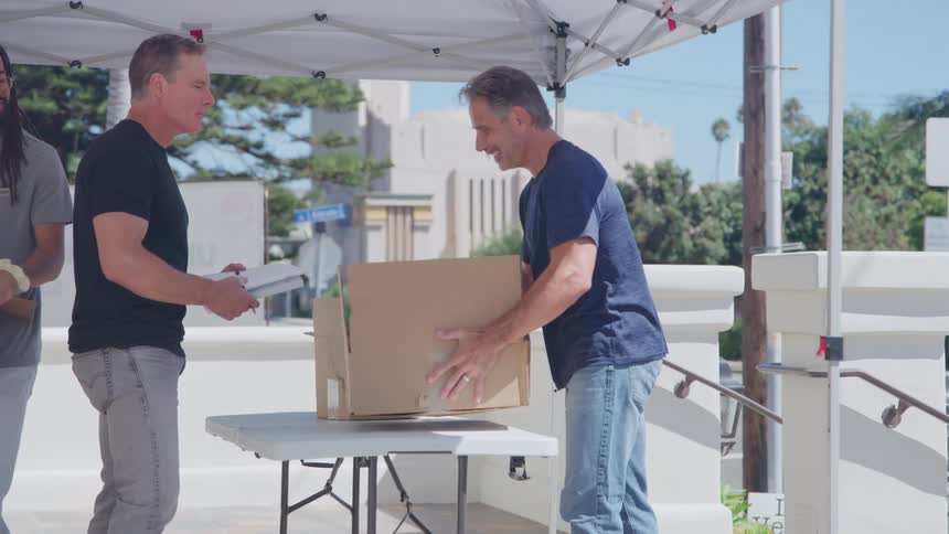 A middleaged man walks up a staircase carrying a cardboard box toward another man standing behind a desk he places the box on the desk exchanges pleasantries while a third man exits the scene holding a cardboard box at midday