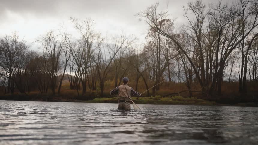 A person fly fishes in a wide river surrounded by bare trees under a cloudy sky